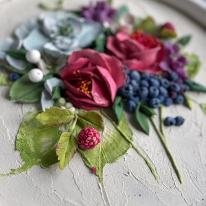 May include: A close-up of a floral artwork with deep red roses, blue berries, and green leaves. The piece has a textured white background, highlighting the vibrant colours and detailed textures of the flowers and foliage.