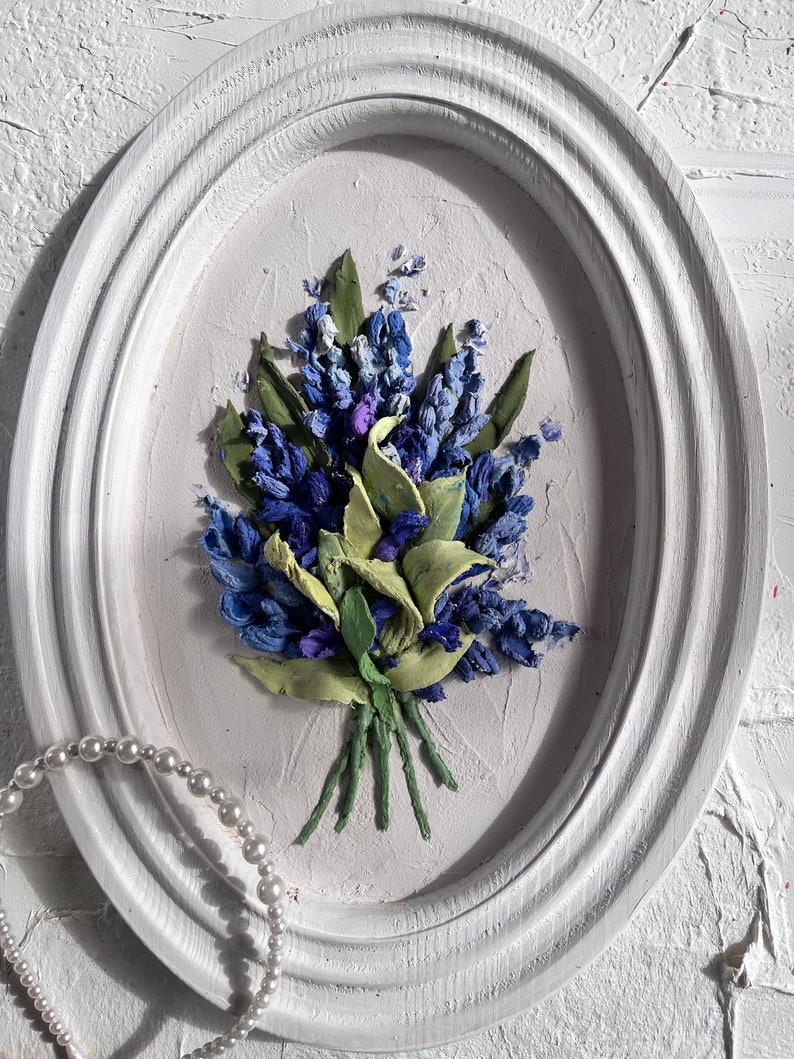 May include: A white oval frame containing a blue and green floral painting. The painting shows a bouquet of blue flowers with green leaves.