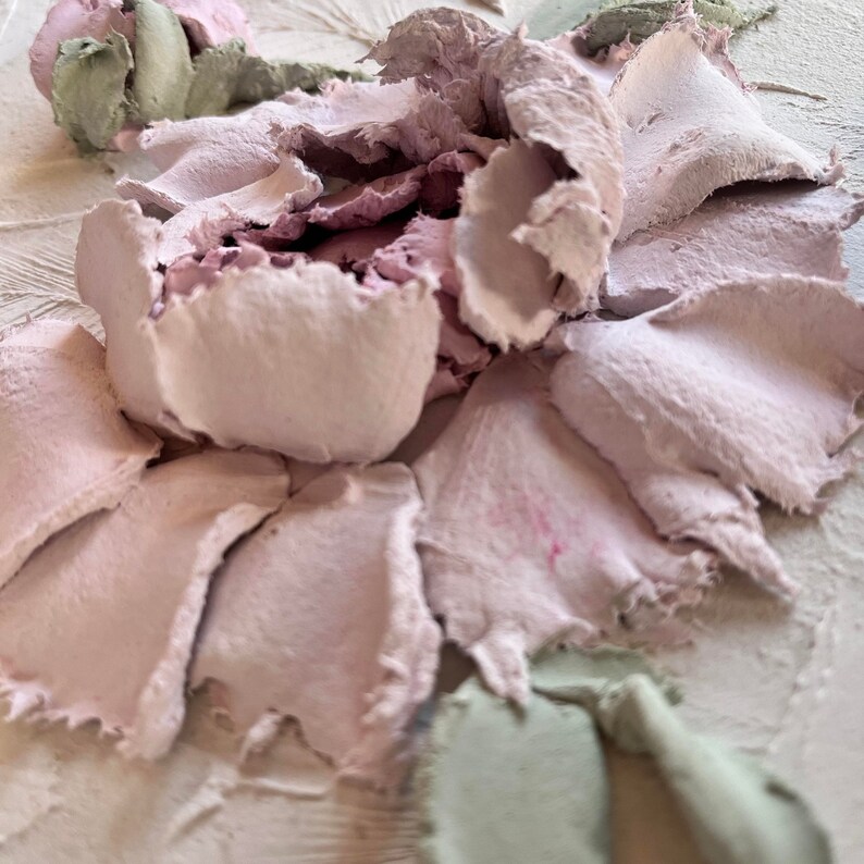 May include: Close-up of a textured artwork featuring a flower design. The petals are a soft pink, with some areas showing darker pink accents. Green and light pink elements are also visible, creating a three-dimensional effect. The background is a light beige.