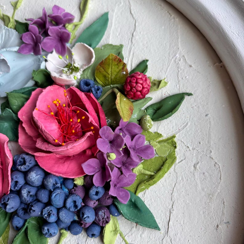 May include: A detailed view of a floral relief artwork. It showcases a pink rose, purple hydrangeas, blueberries, and a red raspberry. The sculpted elements are set against a textured, off-white backdrop, creating a three-dimensional effect.
