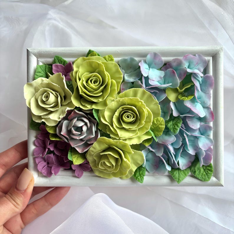 May include: A rectangular white framed floral arrangement featuring green roses, blue-green hydrangeas, and purple accents. The flowers are arranged in a dense, textured composition, creating a three-dimensional effect. The overall aesthetic is delicate and artistic.