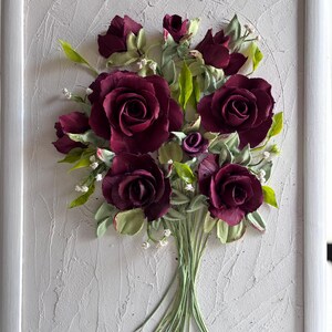 May include: A framed artwork featuring a bouquet of burgundy roses with green leaves and stems. The roses are the focal point, set against a textured white background within a white frame. Small white flowers accent the arrangement.