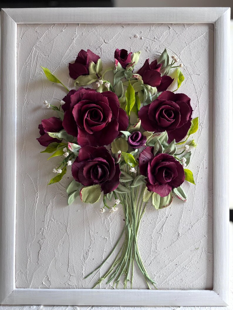 May include: A framed artwork featuring a bouquet of burgundy roses with green leaves and stems. The roses are the focal point, set against a textured white background within a white frame. Small white flowers accent the arrangement.
