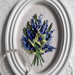 May include: A white oval frame with a blue and green floral embroidery design. The embroidery is of a bouquet of flowers with green stems and leaves. A white pearl headband is in the foreground.