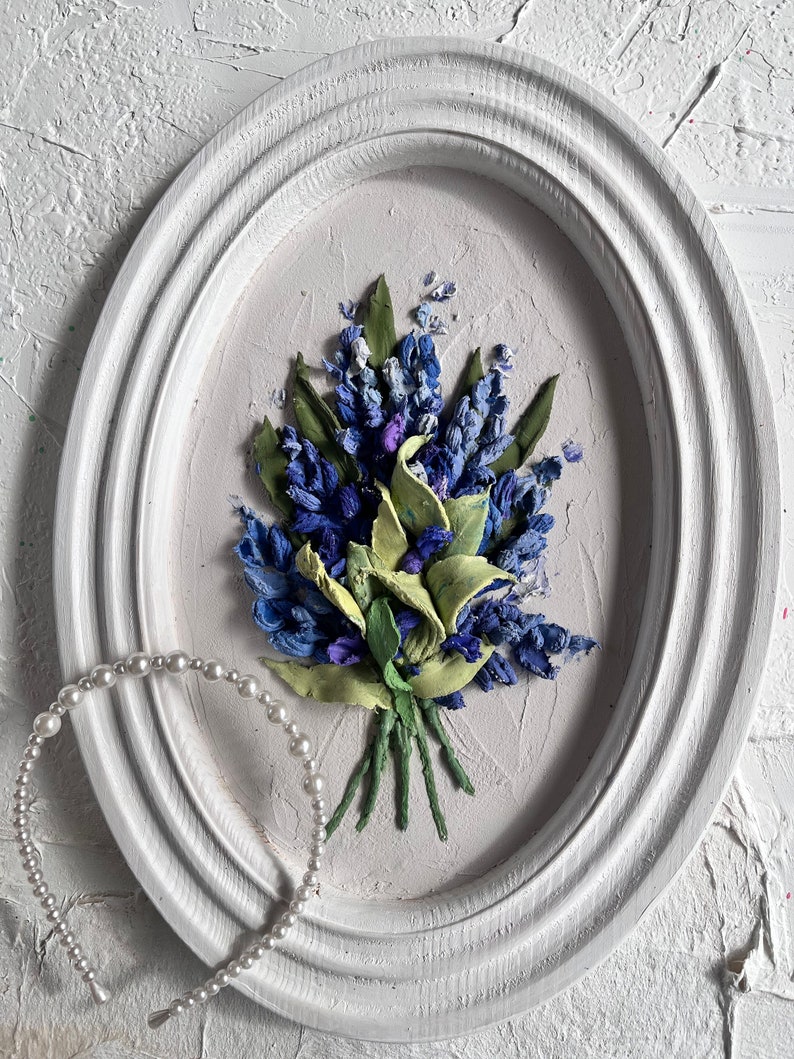 May include: A white oval frame with a blue and green floral embroidery design. The embroidery is of a bouquet of flowers with green stems and leaves. A white pearl headband is in the foreground.