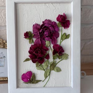 May include: A framed floral artwork featuring deep burgundy flowers with green leaves against a textured white background. The flowers are arranged in a naturalistic style within a white wooden frame, creating a three-dimensional effect. The artwork is a decorative piece.