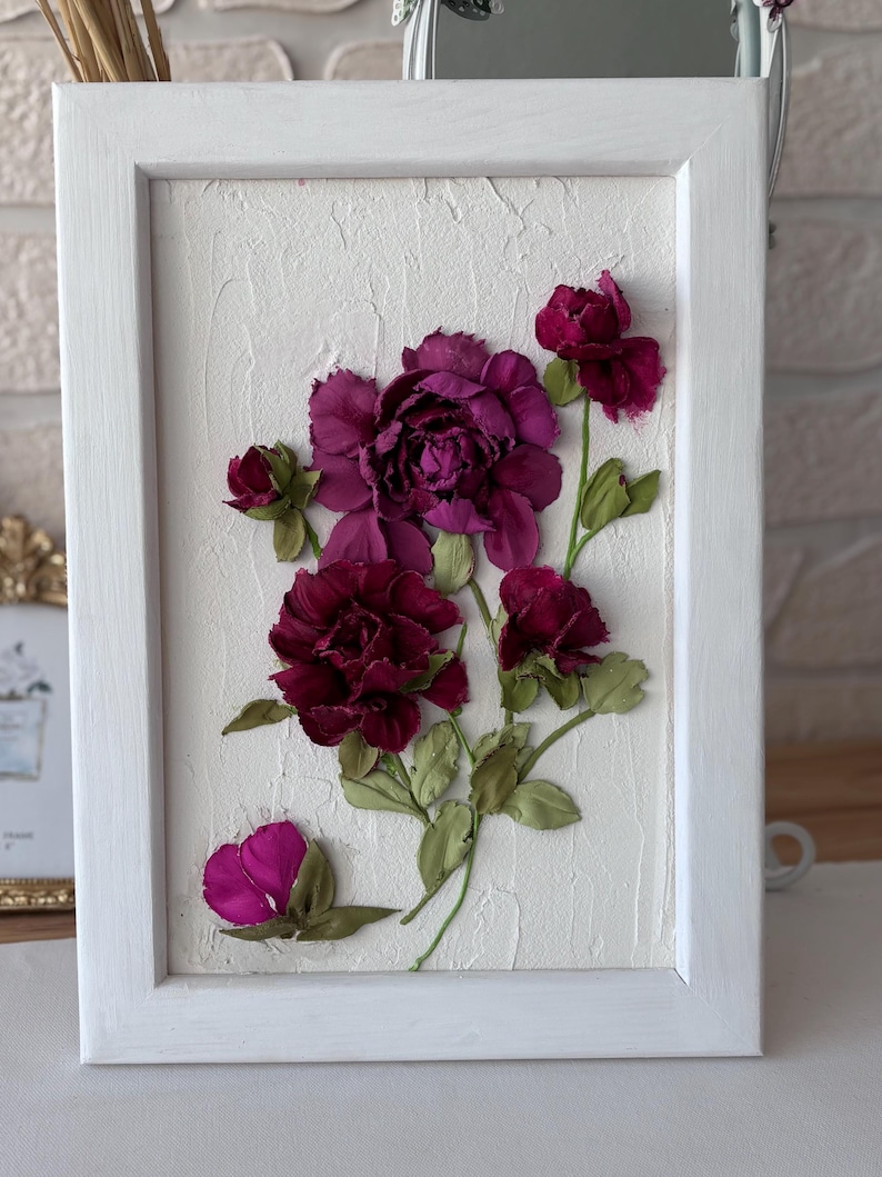 May include: A framed floral artwork featuring deep burgundy flowers with green leaves against a textured white background. The flowers are arranged in a naturalistic style within a white wooden frame, creating a three-dimensional effect. The artwork is a decorative piece.