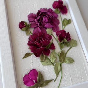 May include: A framed floral artwork featuring burgundy and magenta flowers with green leaves. The flowers are arranged on a textured white background within a white wooden frame. The artwork has a three-dimensional appearance.