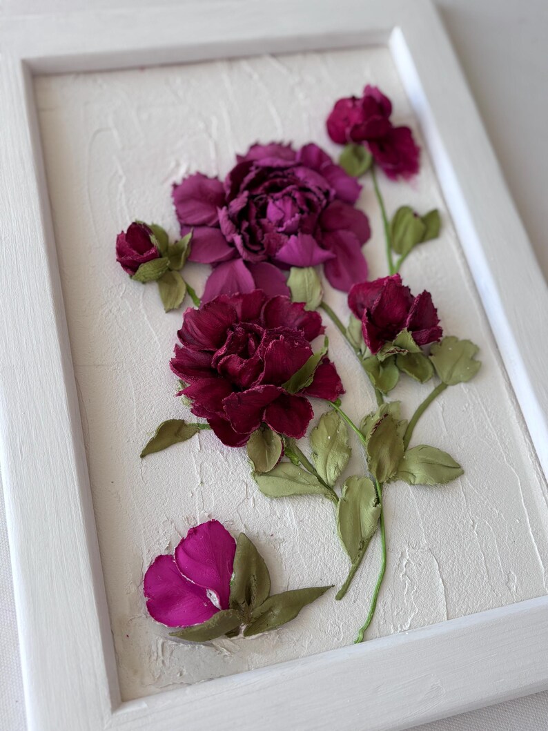 May include: A framed floral artwork featuring burgundy and magenta flowers with green leaves. The flowers are arranged on a textured white background within a white wooden frame. The artwork has a three-dimensional appearance.