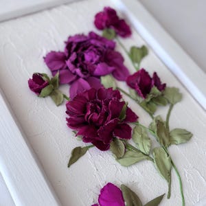 May include: A framed floral artwork featuring dimensional purple flowers and green leaves against a textured white background. The flowers are in various stages of bloom, with a detailed, realistic appearance. The frame is white.