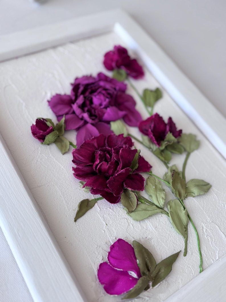 May include: A framed floral artwork featuring dimensional purple flowers and green leaves against a textured white background. The flowers are in various stages of bloom, with a detailed, realistic appearance. The frame is white.