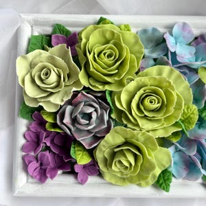 May include: A floral arrangement featuring large, lime green roses, purple hydrangeas, and smaller purple flowers. The flowers are set in a white, rectangular frame, creating a 3D effect. Green leaves are interspersed throughout the arrangement.