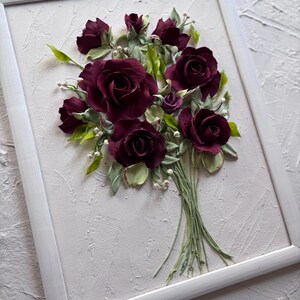 May include: A framed artwork featuring a bouquet of burgundy roses. The roses are crafted with detailed petals and surrounded by green leaves and small white accents. The artwork is set against a textured, off-white background within a white frame.