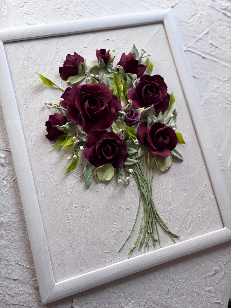 May include: A framed artwork featuring a bouquet of burgundy roses. The roses are crafted with detailed petals and surrounded by green leaves and small white accents. The artwork is set against a textured, off-white background within a white frame.