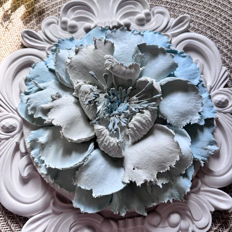 以下が含まれることがあります： 白と薄い青の3Dの花で、テクスチャのあるレイヤーデザインになっています。花は白い装飾的なフレームで囲まれています。