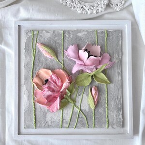 May include: A framed floral artwork featuring textured, three-dimensional pink and peach flowers with green leaves and stems. The flowers are set against a gray background with vertical green lines, all within a white frame.
