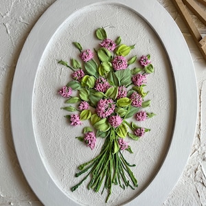 May include: A white oval frame with a white background and a bouquet of pink and green clover flowers. The flowers are made of fabric and are arranged in a realistic style.