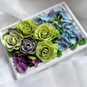 May include: A framed floral arrangement featuring green roses, blue hydrangeas, and purple flowers. The flowers are arranged in a white rectangular frame. The roses are a vibrant green, while the hydrangeas transition from blue to purple.