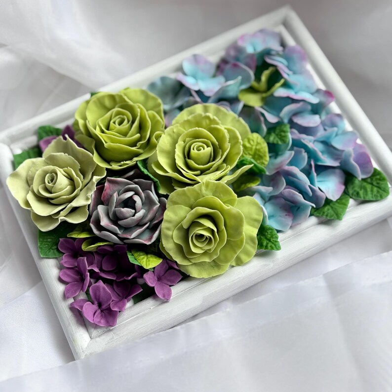 May include: A framed floral arrangement featuring green roses, blue hydrangeas, and purple flowers. The flowers are arranged in a white rectangular frame. The roses are a vibrant green, while the hydrangeas transition from blue to purple.
