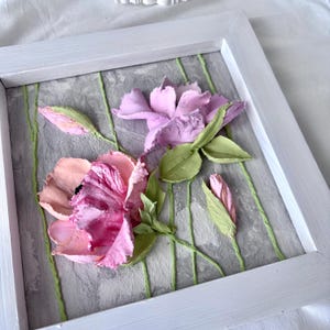 May include: A framed floral artwork featuring textured, three-dimensional flowers in shades of pink and purple. The flowers are set against a gray background with green vertical lines, all within a white wooden frame. The artwork evokes a sense of spring.
