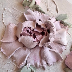 May include: Close-up of a textured, three-dimensional artwork featuring a large, light pink peony flower. The petals have a rough, layered appearance, with a darker pink center. Green leaves and a light beige background complete the floral design.