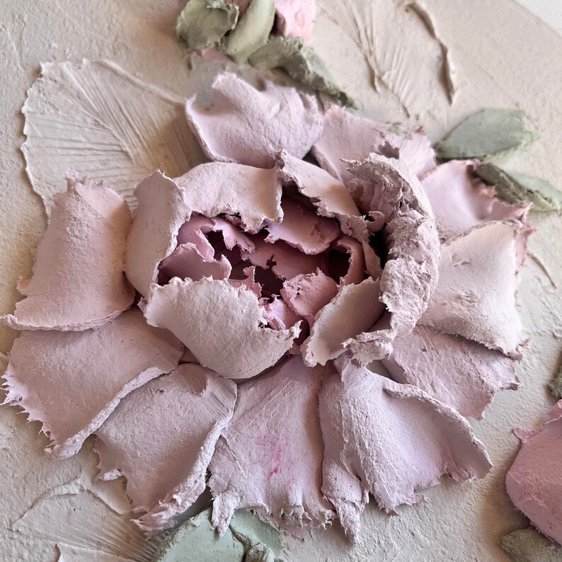 May include: Close-up of a textured, three-dimensional artwork featuring a large, light pink peony flower. The petals have a rough, layered appearance, with a darker pink center. Green leaves and a light beige background complete the floral design.