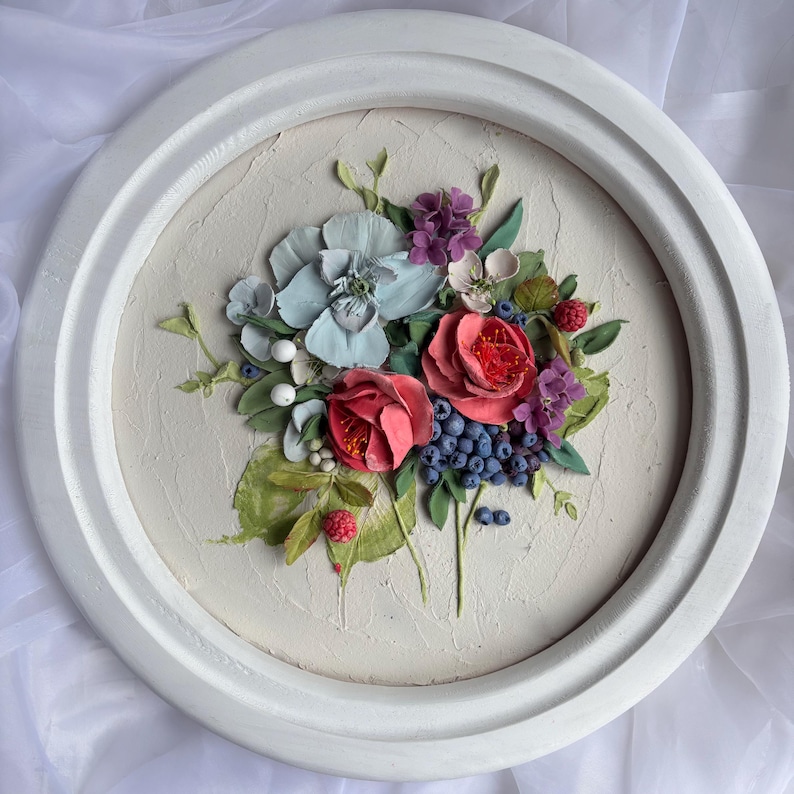 May include: A circular, white-framed relief sculpture of a floral arrangement. The artwork depicts red roses, blue hydrangeas, purple flowers, and blueberries. Green leaves and white berries are set against a textured, off-white background, within a white frame.