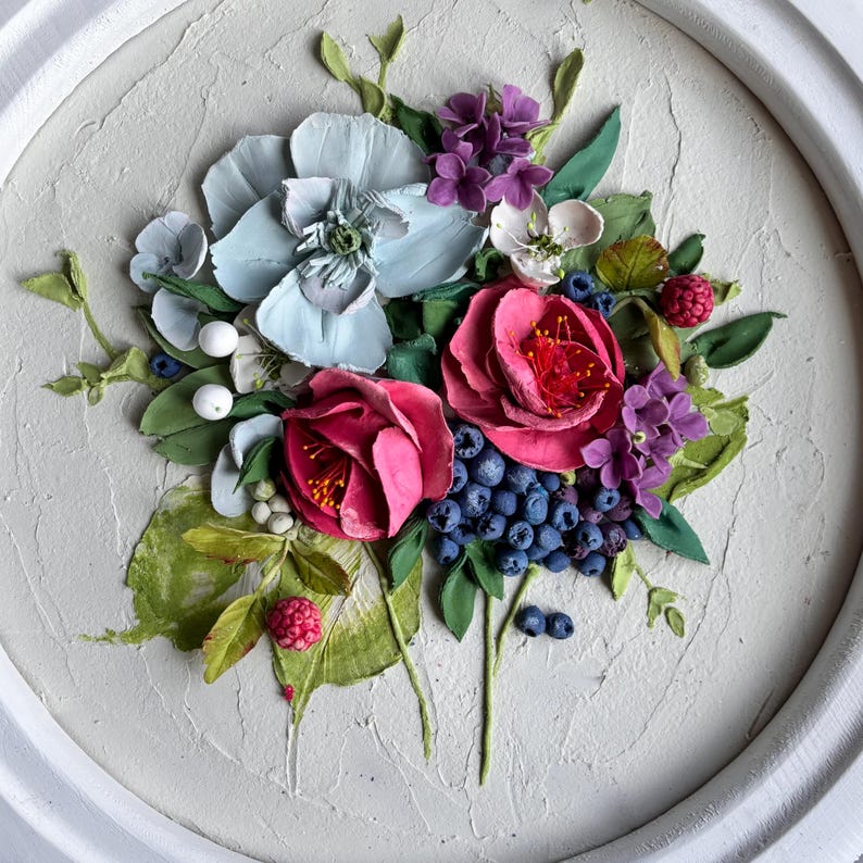 May include: A textured floral artwork showing a bouquet of sculpted flowers and berries. The arrangement includes large pink roses, blue-grey blossoms, purple hydrangeas, and blueberries, with green leaves and white berries. The artwork is set in a white, circular frame.