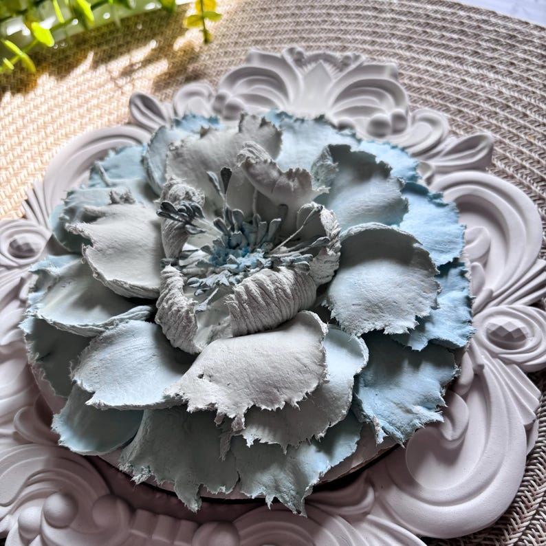 以下が含まれることがあります： 白と薄い青色の紙の花で、表面にテクスチャがあります。花は白い装飾的なフレームに入っています。