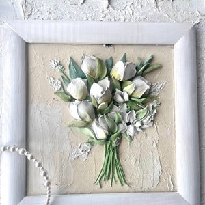 May include: A framed white and green 3D floral painting of a bouquet of white flowers with green leaves. The painting is on a beige background and is framed in a white frame.