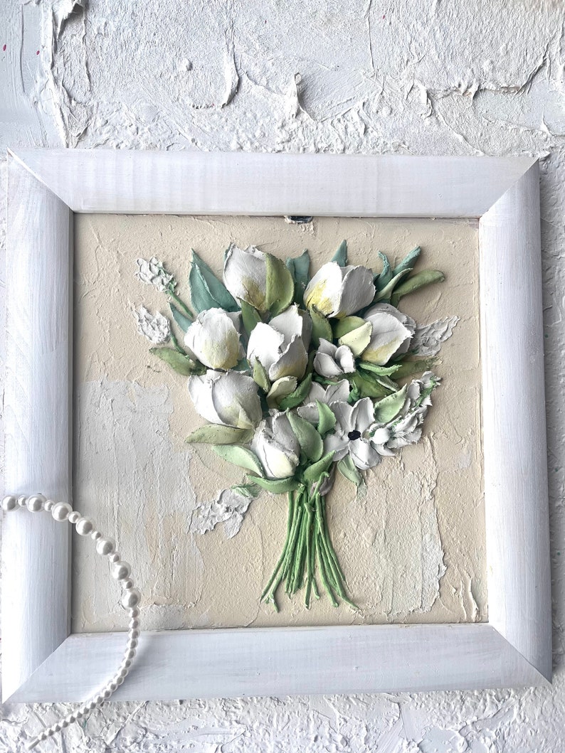 May include: A framed white and green 3D floral painting of a bouquet of white flowers with green leaves. The painting is on a beige background and is framed in a white frame.