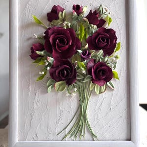 May include: A framed floral artwork featuring a bouquet of deep burgundy roses with green leaves and small white flowers. The flowers are arranged in a bouquet and set against a textured, off-white background within a white frame.
