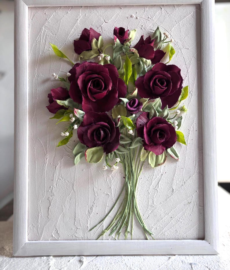 May include: A framed floral artwork featuring a bouquet of deep burgundy roses with green leaves and small white flowers. The flowers are arranged in a bouquet and set against a textured, off-white background within a white frame.