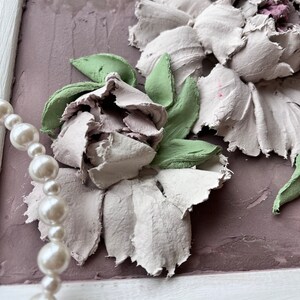 May include: A framed artwork featuring a 3D floral design with white and green flowers. The flowers are made of textured paper or fabric and are arranged in a cluster. The background is a light purple color.