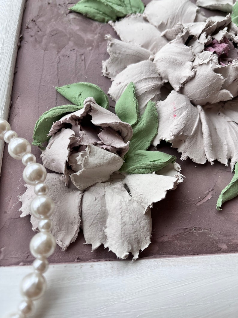 May include: A framed artwork featuring a 3D floral design with white and green flowers. The flowers are made of textured paper or fabric and are arranged in a cluster. The background is a light purple color.