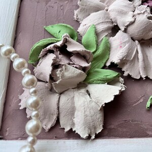 May include: A close-up of a white and green floral painting with a pearl necklace in the foreground. The painting is framed in white and has a textured background.