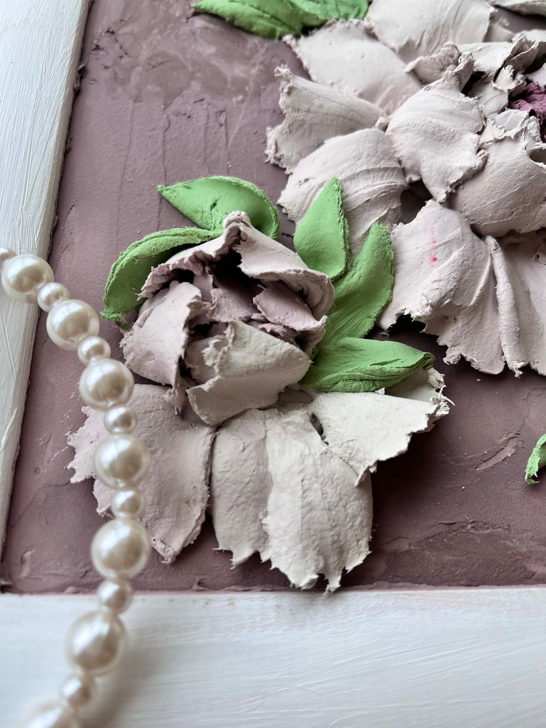 May include: A close-up of a white and green floral painting with a pearl necklace in the foreground. The painting is framed in white and has a textured background.