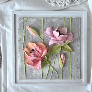 May include: A framed floral artwork featuring textured, three-dimensional flowers in shades of pink and peach. Green stems and leaves accent the blossoms against a gray background with vertical green lines. The artwork is encased in a white frame.