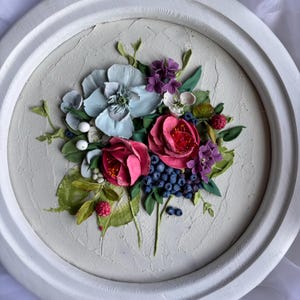 May include: A circular, white-framed relief sculpture of a floral arrangement. The artwork depicts sculpted flowers in pink, blue, and purple, with green leaves and blue berries. The background has a textured, off-white finish.