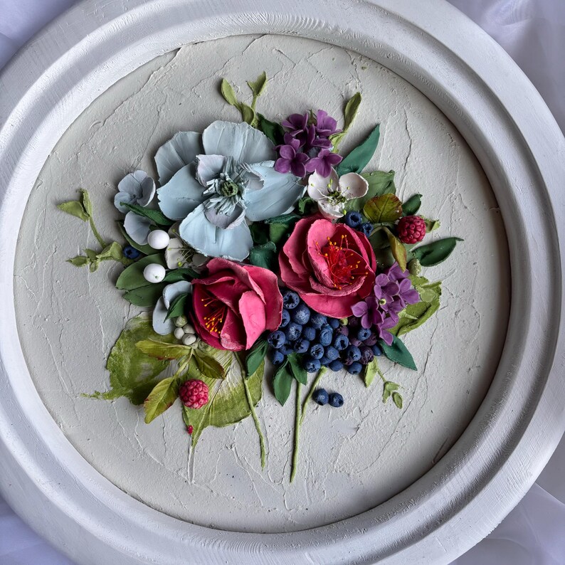 May include: A circular, white-framed relief sculpture of a floral arrangement. The artwork depicts sculpted flowers in pink, blue, and purple, with green leaves and blue berries. The background has a textured, off-white finish.
