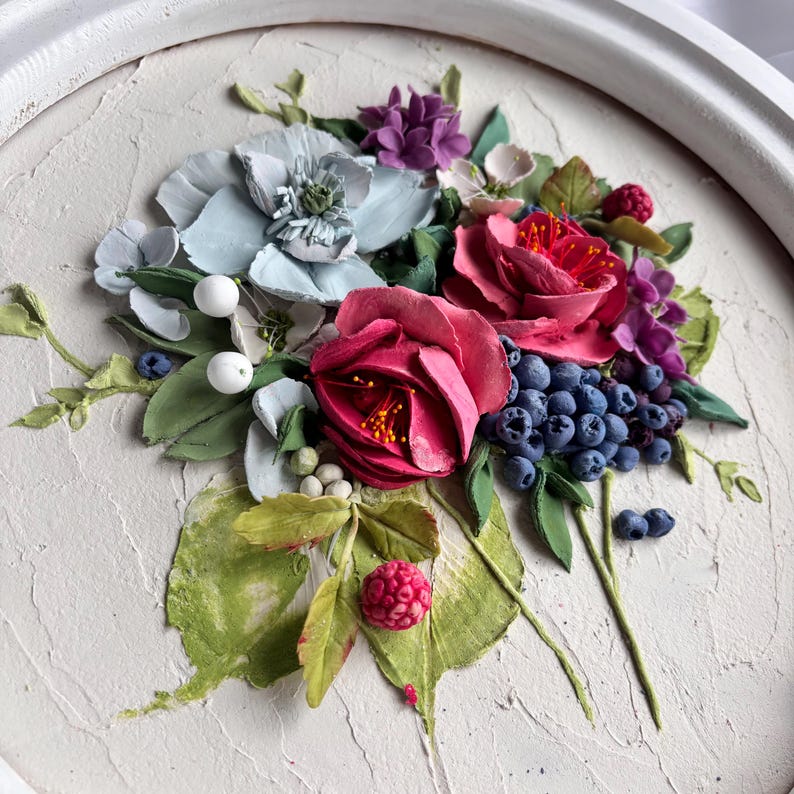 May include: A detailed artwork featuring a bouquet of sculpted flowers and berries. The arrangement includes deep red roses, light blue blossoms, purple hydrangeas, and clusters of blueberries and white berries. The piece is set against a textured, off-white background within a white frame.
