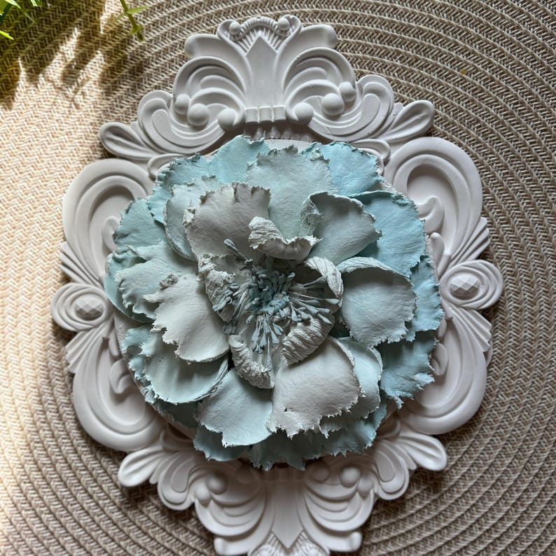 以下が含まれることがあります： 白い装飾的なフレームの中央に、薄い青と白の紙の花が飾られています。
