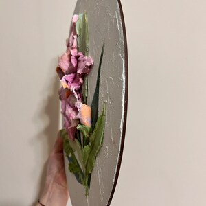 K&ouml;nnte beinhalten: Ein rundes, strukturiertes Leinwandbild einer rosa und orangefarbenen Blume mit gr&uuml;nen Bl&auml;ttern. Das Bild ist auf einem Holzrahmen.
