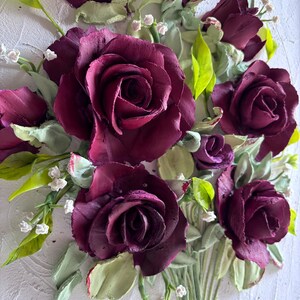 May include: A detailed, three-dimensional floral artwork featuring deep burgundy roses. The roses are the focal point, surrounded by green leaves and small white flowers. The artwork is set against a textured white background, creating a striking contrast.