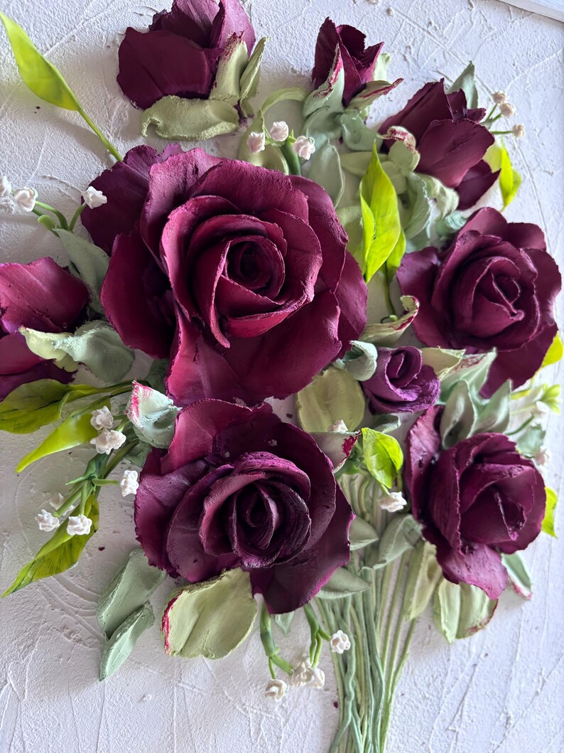May include: A detailed, three-dimensional floral artwork featuring deep burgundy roses. The roses are the focal point, surrounded by green leaves and small white flowers. The artwork is set against a textured white background, creating a striking contrast.