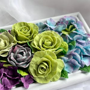 May include: A floral arrangement featuring handcrafted roses, hydrangeas, and other flowers in a white frame. The roses are a vibrant green, with purple, blue, and green accents. The flowers are arranged in a rectangular shape.