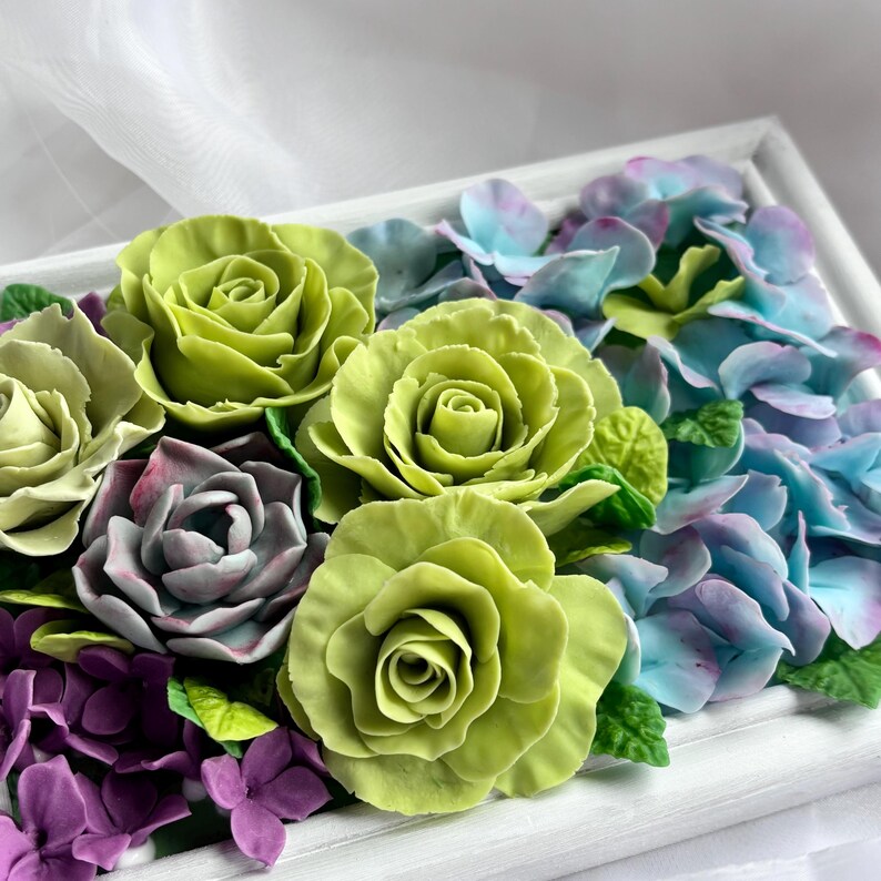 May include: A floral arrangement featuring handcrafted roses, hydrangeas, and other flowers in a white frame. The roses are a vibrant green, with purple, blue, and green accents. The flowers are arranged in a rectangular shape.