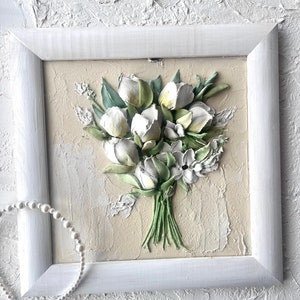 May include: A white framed picture with a 3D textured painting of a bouquet of white flowers with green leaves on a beige background.