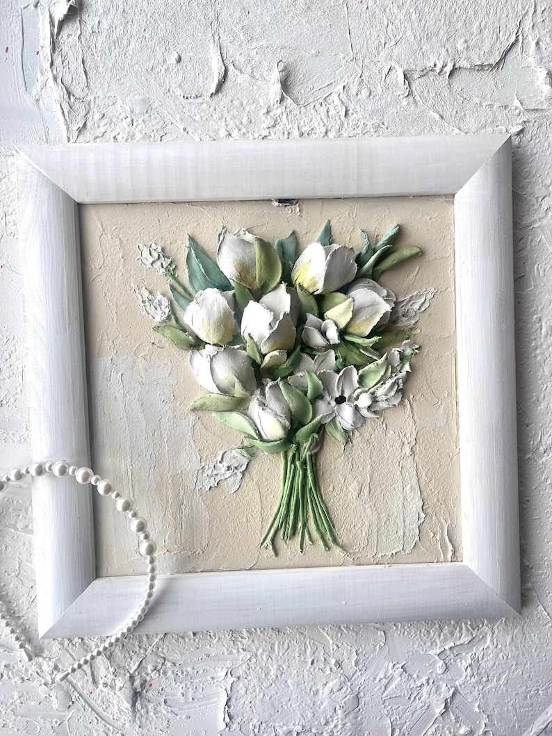 May include: A white framed picture with a 3D textured painting of a bouquet of white flowers with green leaves on a beige background.