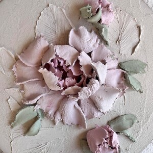 May include: A textured, round, off-white artwork featuring a detailed, raised floral design. The central flower is a soft pink, surrounded by textured petals. Green leaves and smaller pink buds accent the design, creating a three-dimensional effect.